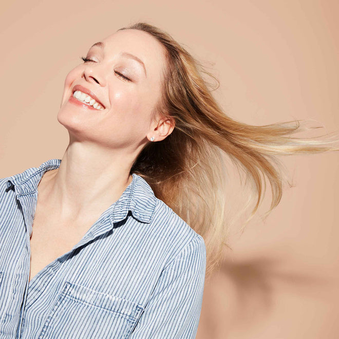 A smiling woman, eyes closed, tilts her head back and tosses her long, blonde hair over her shoulder. She wears a blue and white striped shirt against a plain light orange background.