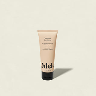 A tube of Volumizing Conditioner Mini, a salon-grade formula for weightless moisture, stands upright on a light beige background. The peach-colored tube has black text and a black cap, casting a soft shadow.