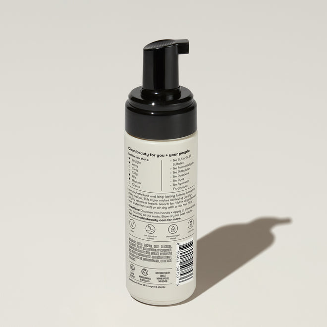 A bottle of Odele Volumizing Foam stands upright against a plain light-colored backdrop with the back of the label facing the camera.
