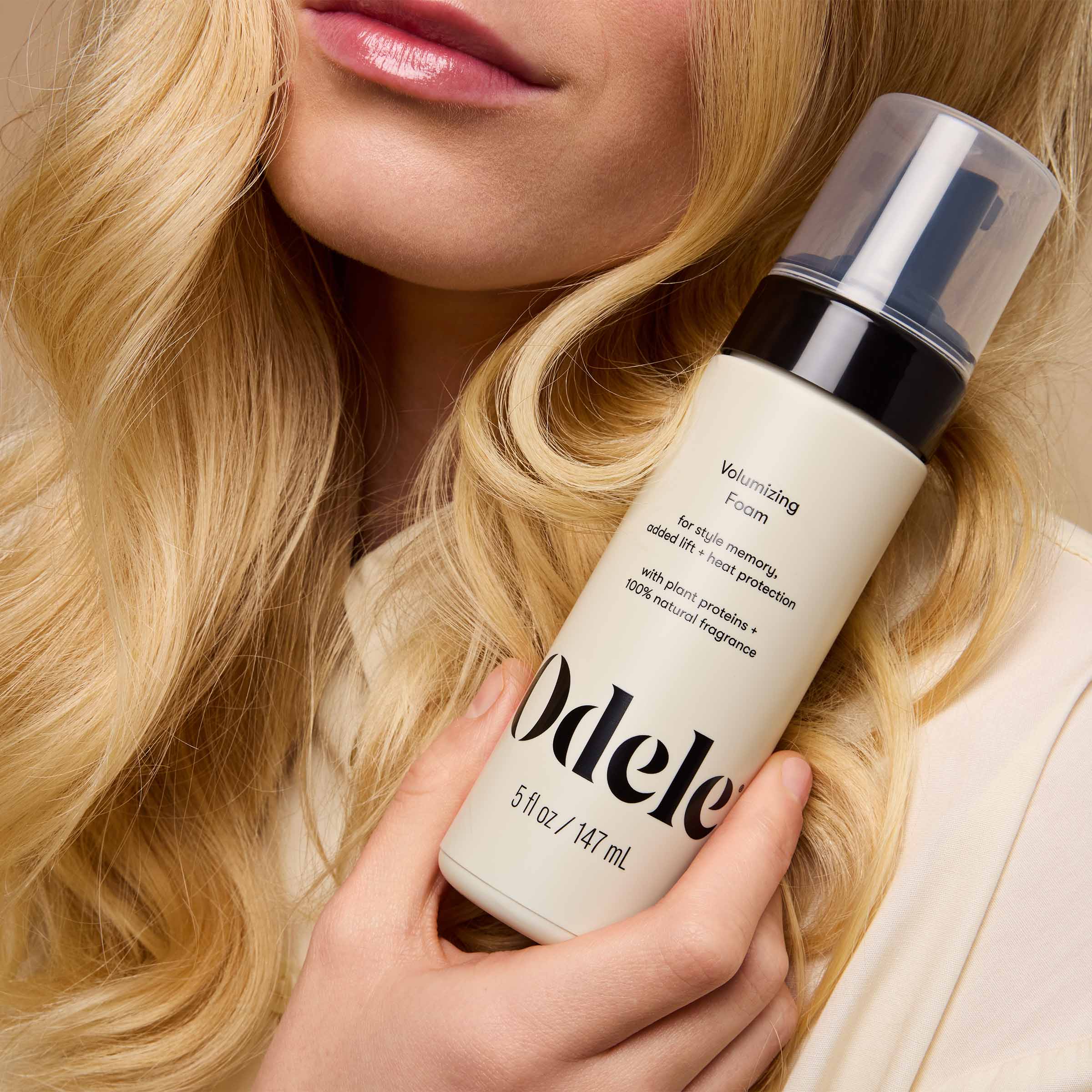 A woman with long, wavy blonde hair holds a bottle of Odele Volumizing Foam near her face.