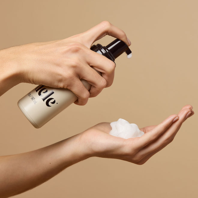 A person dispenses Odele Volumizing Foam onto their palm against a neutral beige background.