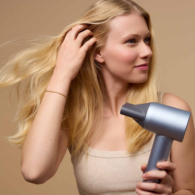 A young woman uses a silver blow dryer on her long blonde hair. She wears a beige tank top and looks to the side against a neutral background.