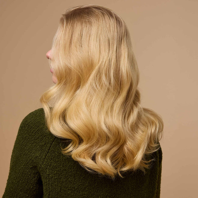 A woman with long, wavy blonde hair is shown from behind in a textured dark green sweater against a plain beige background.