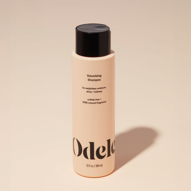A bottle of Odele Volumizing Shampoo stands upright against a plain, light orange backdrop with the front of the label facing the camera.