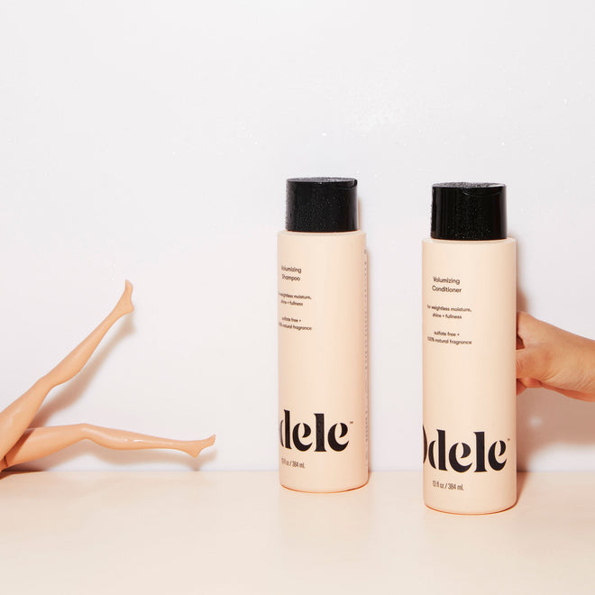 Bottles of Odele Volumizing Shampoo and Conditioner stand upright on a white surface. A hand holds one bottle, while two doll legs are visible on the left against a plain white background.