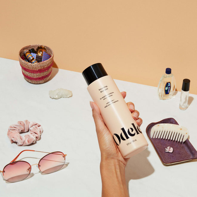 A hand holds a bottle of Odele Volumizing Shampoo above a white surface scattered with miscellaneous accessories.
