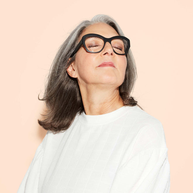 A confident older woman with gray hair and glasses, wearing a white top, poses with her head tipped upwards and eyes closed against a light peach background.