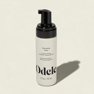A white bottle of Volumizing Foam with a black pump rests on a light background, casting a shadow. The label notes plant proteins, natural fragrance, heat protection, and the size: 5 fl oz (147 mL).