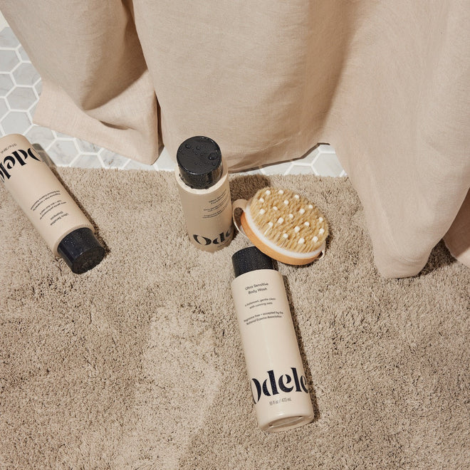Three beige bottles from the Odele Beauty Ultra Sensitive Set lay on a beige shower mat with a wooden body brush.