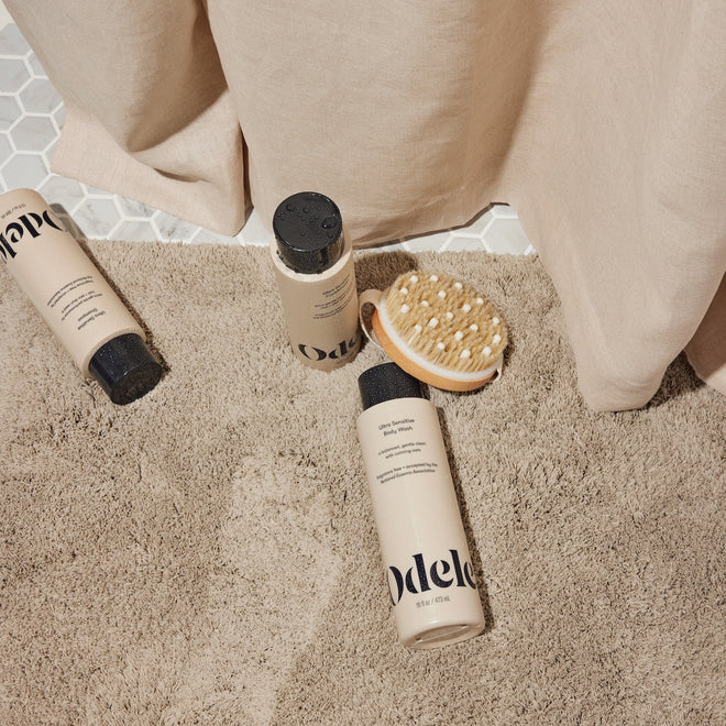 Three beige bottles from the Odele Beauty Ultra Sensitive Set lay on a beige shower mat with a wooden body brush.