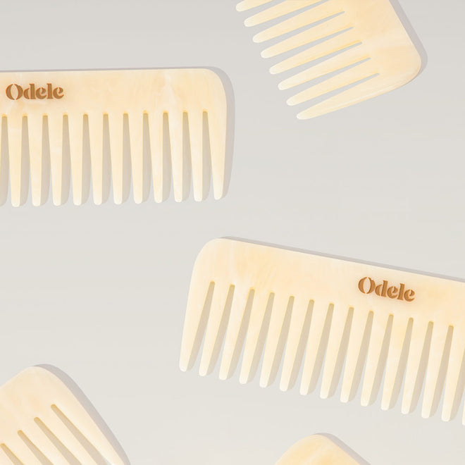 Several light beige wide-toothed combs with a gold Odele logo on each handle lay scattered on a plain, light-colored background.