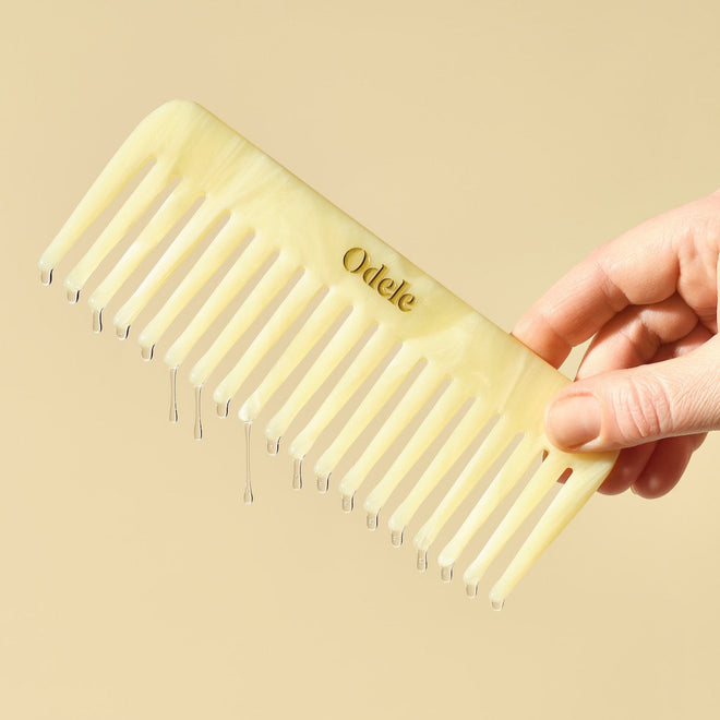 A hand holds a tan wide-toothed comb dripping with oil in front of a plain light orange background.
