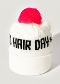 A white knit beanie with a bright pink pom-pom and “Odele” written in black across the front stands upright against a plain white background.