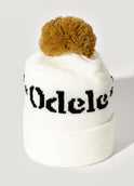 A white knit beanie with a dark gold pom-pom and “Odele” written in black across the front stands upright against a plain white background.